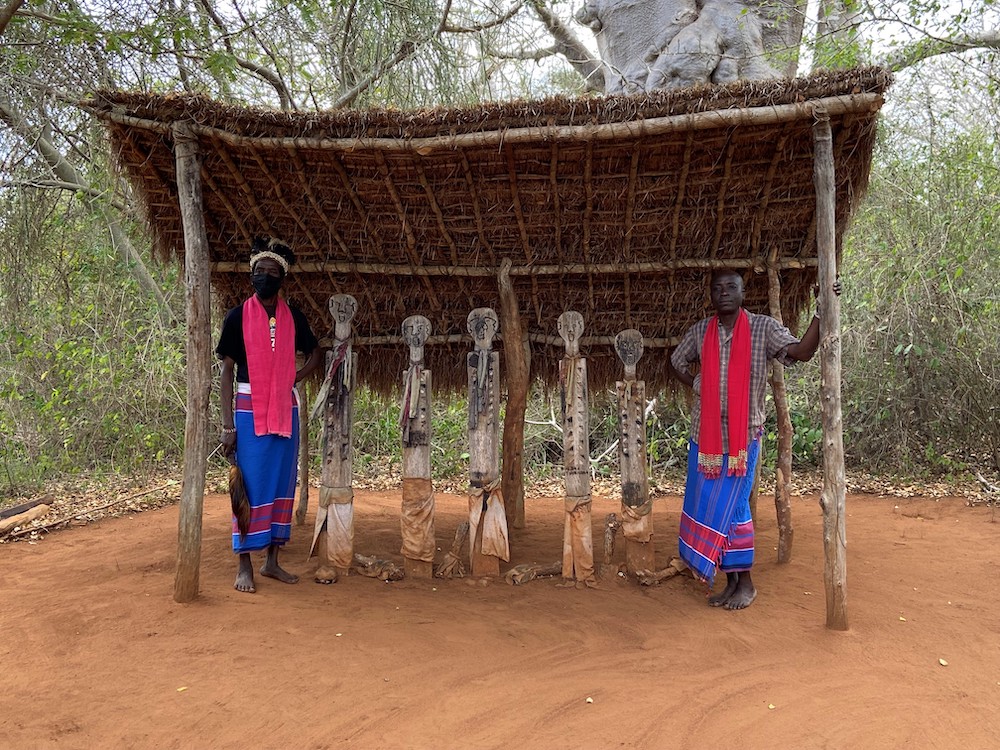Two people wearing red and blue clothing stand on either side of five wooden sculptures under a shelter with a thatched roof.